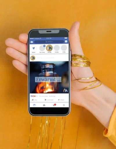 Hand mit goldenen Armreifen hält iPhone mit Facebook-Post zu ‚Erwärmt...‘ – Foto einer brennenden Glaslampe auf gelbem Hintergrund
