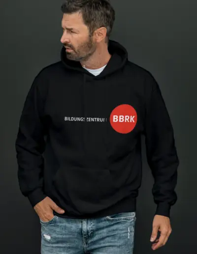 Mann mit Bart in schwarzem Hoodie ‚BILDUNG:ZENTRUM‘ mit rotem Punkt und ‚BBRK‘, lässig mit Händen in den Taschen einer zerrissenen Jeans auf grauem Hintergrund