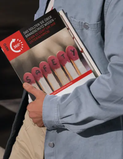 Mann in Hemd und Jeansjacke hält Buch ‚Das Brandschutz-Wissen, das Sie in Ihrem Unternehmen brauchen‘ mit roten Streichhölzern auf Cover, roter Titel ‚360° Infos zum Brandschutz