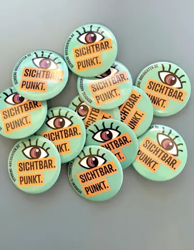Stapel bunter Buttons mit Augen-Motiv und Texten wie ‚Sichtbar. Punkt.‘ für Schöne Aussichten e.V. auf grauem Hintergrund