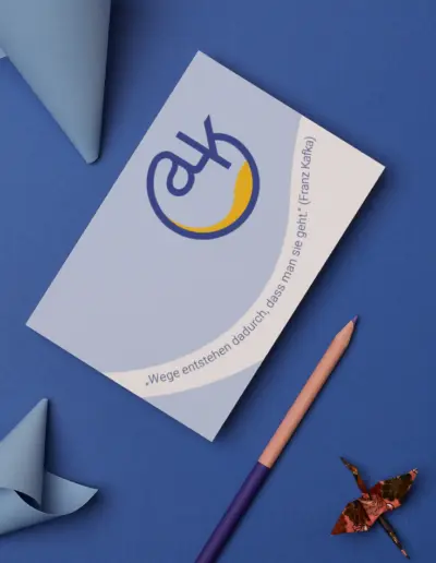 AK: Visitenkarte mit Logo ‚AK‘ in blau-gelber Kurve, Text ‚Wege entstehen durch das Gehen‘ (Franz Kafka), neben Origami-Kranich, Stift und Papier auf blauem Hintergrund